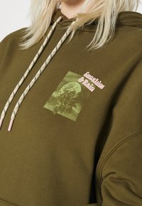 Un sweat à capuche vert olive avec des cordons de tirage blancs et beiges, présentant un graphique rose avec le texte "Soleil et Pluie" et un motif floral.