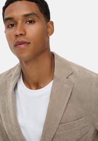 Beiger Cordblazer mit einer einzelnen Tasche, strukturiertem Kragen und weicher Textur, über einem weißen T-Shirt geschichtet.