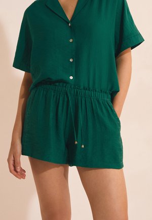 Kobieta ubrana w zielony top z krótkim rękawem i guzikami oraz dopasowane szorty z elastycznym pasem i sznurkami, stojąca z jedną ręką w kieszeni.