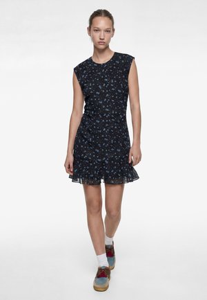 Donna con un vestito nero senza maniche con stampa floreale blu e orlo con volant, calzini bianchi e scarpe multicolori con lacci, cammina in avanti.