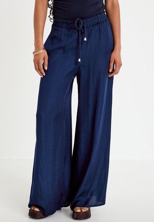 Femme portant un pantalon large bleu marine avec une taille à cordon et des sandales compensées marron, debout sur un fond blanc.
