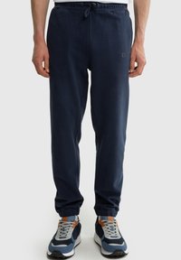 Muž v tmavě modrých teplákových kalhotách s elastickými manžetami a odpovídajícími modro-šedo-oranžovými teniskami stojí před jednoduchým pozadím.