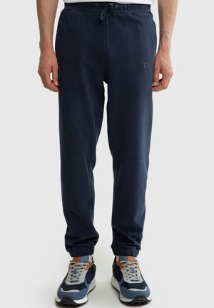 Mężczyzna noszący granatowe spodnie dresowe ze ściągaczami u dołu i pasujące niebiesko-szaro-pomarańczowe sneakersy, stojący na tle jednolitego tła.