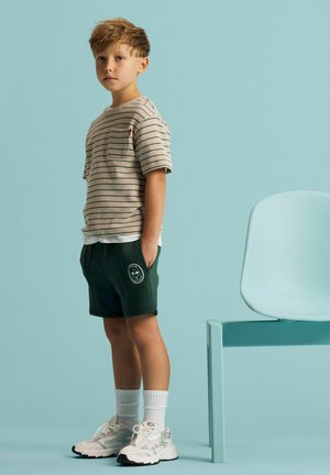 Ung dreng iført en beige t-shirt med striber, mørke shorts, hvide sokker og sneakers står ved siden af en lyseblå stol mod en blå baggrund.