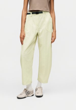 Personne portant un pantalon court beige clair avec de grandes poches avant, une ceinture noire, des chaussettes blanches et des baskets beiges avec des détails noirs et des chiffres.