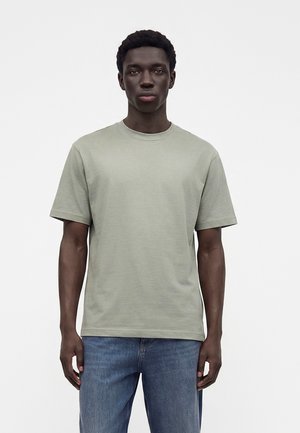 Jeune homme à la peau foncée et aux cheveux courts et bouclés, portant un t-shirt gris clair à manches courtes et un jean bleu, debout devant un fond blanc.