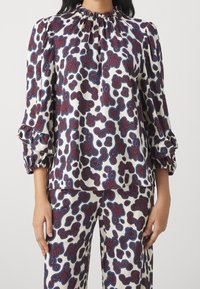 Blusa stampata con collo alto e maniche arricciate, di colore crema con motivi astratti in bordeaux e blu. Tessuto leggero e morbido.