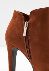 Stivale ankle in suede marrone con tacco a stiletto, dotato di cerniera laterale e dettagli cuciti. Texture liscia e design elegante.