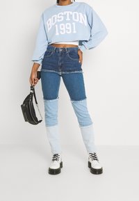 Sudadera recortada de color azul claro con "BOSTON 1991" en blanco, combinada con jeans de mezclilla de parcheo que presentan diferentes tonalidades de azul y bordes deshilachados.