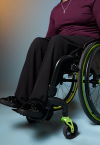 Μαύρο αναπηρικό καροτσάκι με πράσινα αξεσουάρ, φορεμένο με φαρδιά μαύρα παντελόνια και γυαλιστερά μαύρα αθλητικά παπούτσια. Λείες υφές και σύγχρονες λεπτομέρειες σχεδίασης.