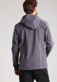 Homme portant une veste à capuche grise avec des coutures verticales jaunes, debout de dos sur un fond gris clair uni.