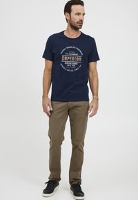 Marineblauwe katoenen T-shirt met een bedrukte ronde logo in beige en witte tekst, gecombineerd met khaki broek en zwarte sneakers.
