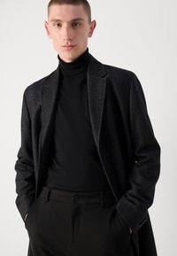 Cappotto strutturato nero con una vestibilità su misura, indossato sopra un dolcevita nero. Presenta rever eleganti e decorazioni con motivi sottili.