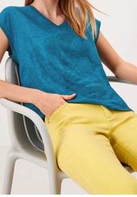 Blusa de lino en color azul verdecido con escote en forma de V y patrón de hojas, combinada con pantalones amarillos. La blusa cuenta con mangas cortas y un ajuste relajado.