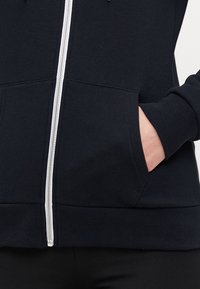 Marineblå hettejakke med hvit glidelås, ribbete mansjetter og hem, samt frontlommer. Stoffet ser mykt ut med en glatt tekstur.