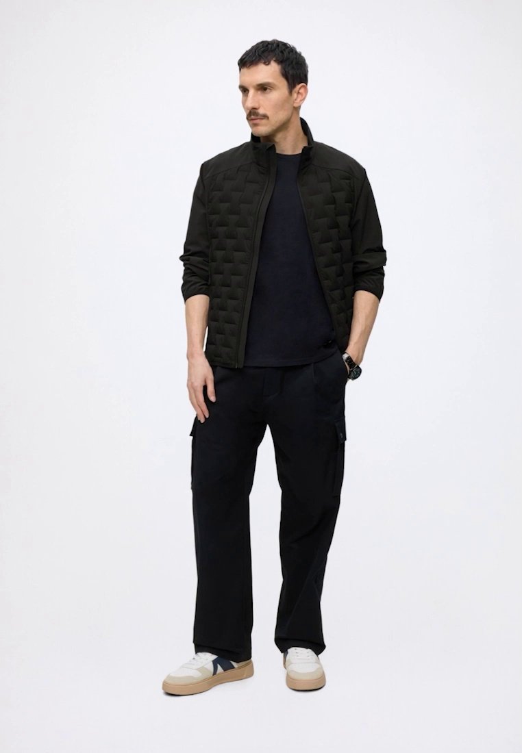 Homme aux cheveux courts et foncés et à la moustache, portant une veste matelassée noire, un pantalon noir et des baskets blanches à semelles beiges, debout les mains dans les poches.