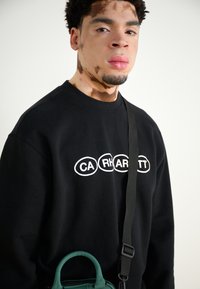 Sudadera negra con un logotipo blanco en forma ovalada. Combinada con un pequeño bolso verde con un asa redondeada y una correa negra.