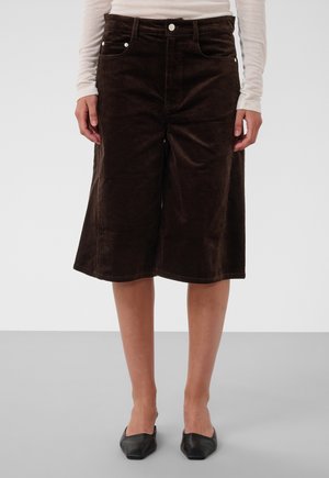 Shorts en velours côtelé marron jusqu'aux genoux, à jambes larges, portés avec un haut blanc à manches longues et des ballerines noires à bout pointu sur un fond clair.