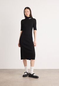 Abito midi a maglia nera a coste con maniche corte, caratterizzato da un colletto alto e una fessura laterale. Abbinato a scarpe nere e calzini bianchi.