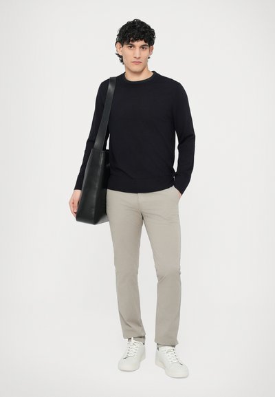 Czarny sweter z prążkowanym dekoltem, beżowe chinosy i białe sneakersy. Modelka niesie czarną skórzaną torbę typu tote, stoi na tle białego tła.