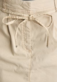 Beige rövidnadrág, állítható derékrésszel, fémből készült végződésekkel, elülső redőkkel és kontrasztos varrási részletekkel a derékrészen.