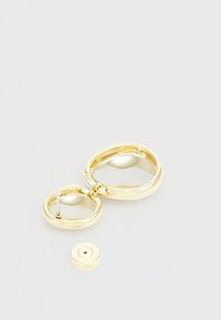 Boucles d'oreilles en ton or avec un design lisse et arrondi, finition polie ; comprend un petit dos magnétique rond avec le nom de la marque.