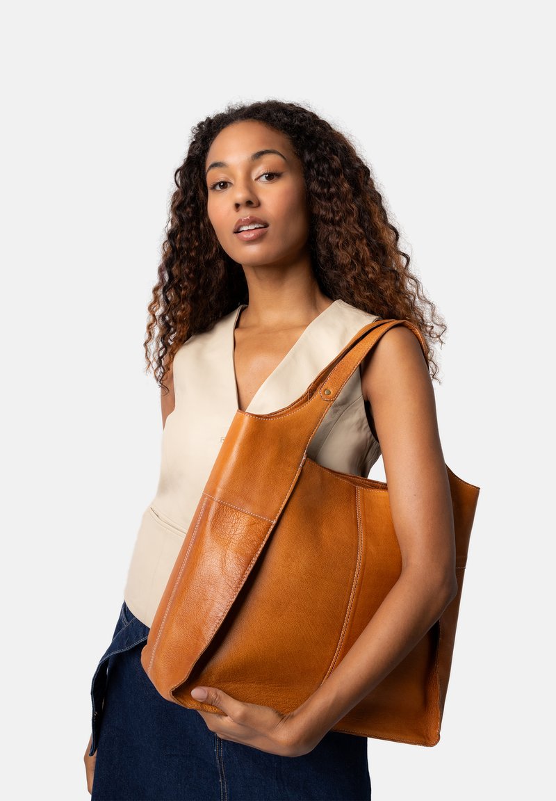 Sac fourre-tout en cuir marron avec une texture lisse, de grandes anses, et un détail de surpiqûre minimal, tenu par un modèle portant un haut beige.
