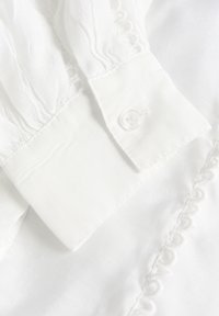 Weißes Baumwollblusenkleid mit Rüschenmanschetten, versehen mit einem runden Knopf und zarter Stickerei entlang der Naht. Weicher Stoff, leichtes Material.