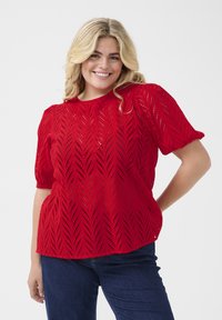 Femme souriante aux cheveux blonds portant un haut à motifs rouges à manches courtes et un jean bleu foncé, posant avec une main sur la hanche.