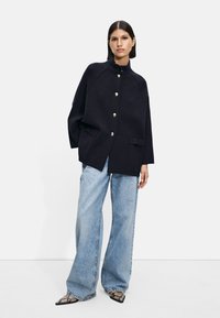 Marineblå oversized jakke med gullknapper, avslappet passform og to frontlommer, kombinert med lyseblå vide jeans og mønstrede sko.