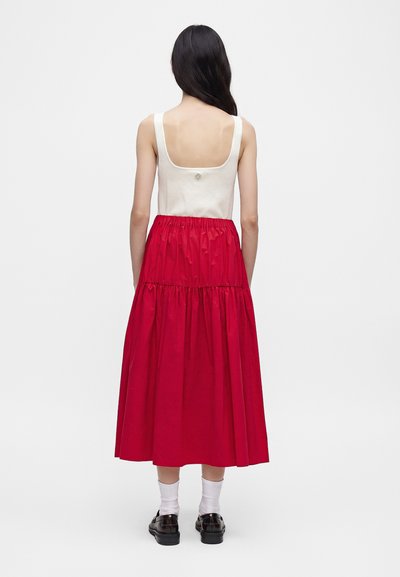 Femme aux longs cheveux foncés portant un haut blanc sans manches et une jupe midi rouge froncée, debout de dos sur un fond uni.