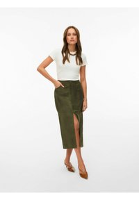 Femme portant un haut blanc ajusté à manches courtes, une jupe midi olive avec une fente à l'avant, des talons pointus marron et un collier chunky, debout avec une main dans la poche.