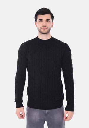 Maglione nero a maglia con motivo a trecce, scollo rotondo e polsini e orlo a costine. Indossato sopra jeans grigi chiari.