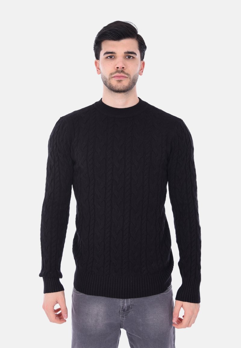 Maglione nero a maglia con motivo a trecce, scollo rotondo e polsini e orlo a costine. Indossato sopra jeans grigi chiari.