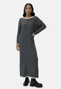 Vestido maxi gris acanalado con ribete blanco en el escote y los puños de las mangas, con mangas largas y bolsillos laterales. Combinado con botas negras de plataforma.