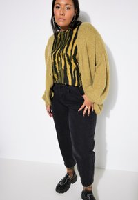 Přehnaně velký žlutozelený pletený cardigan přes černé a žluté tričko s tygrovaným vzorem, doplněný o černé džíny s vysokým pasem a masivní černé boty.