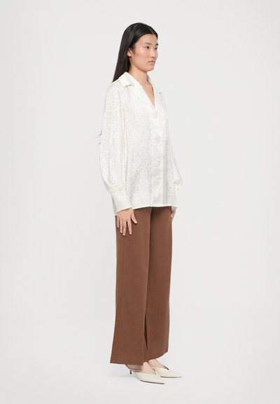 White blouse with textured pattern and puffed sleeves, paired with brown wide-leg trousers and cream-colored heels, shown against a plain background.
