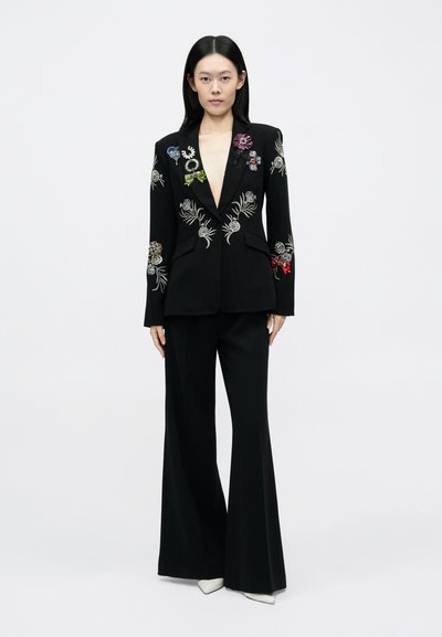 Fekete blézer részletes, színes virágmintás hímzéssel, széles szárú fekete nadrággal és hegyes orrú fehér magassarkúval párosítva.