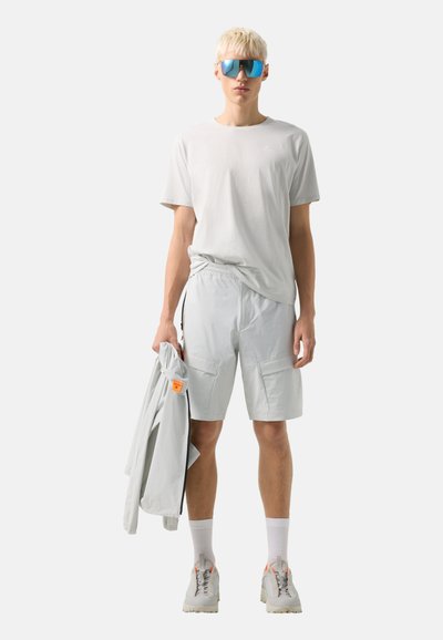 Młoda osoba z krótkimi blond włosami, w okularach przeciwsłonecznych z odblaskowymi soczewkami, jasnoszarym t-shircie, szortach, białych skarpetkach i trampkach, trzymająca jasną kurtkę.