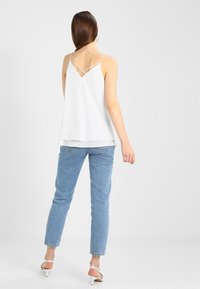 Valkoinen hihaton toppi, jossa on lentävä muotoilu, ohuet olkaimet ja ristiselkäyksityiskohta, yhdistettynä vaaleansinisissä suoriin farkkuihin ja valkoisiin korkokenkiin.