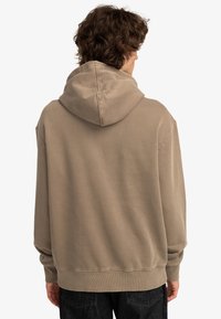 Hoodie i ljusbrun bomull, avslappnad passform, med ribbade ärmslut och nederkant, samt en dubbelstickad huva. Bakåtblick.