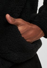 Jersey de forro polar negro con un exterior texturizado y acogedor y un bolsillo tipo canguro; una mano descansando dentro del bolsillo muestra el pulgar.