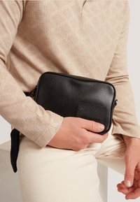 Bolso de mano negro de cuero con una textura suave, cierre de cremallera y un pequeño bolsillo frontal. Sostenido contra un suéter beige con un patrón.