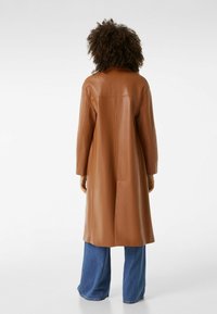 Hnědý kožený kabát s rovný střih, dlouhými rukávy a subtilním prošíváním, doplněný o kalhoty s širokými nohavicemi z denimu. Hladká textura.