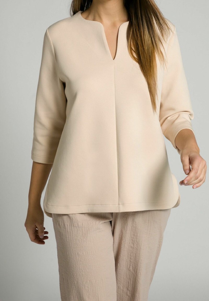 Femme portant un haut ample beige à manches trois-quarts avec col en V et un pantalon beige texturé assorti sur fond neutre.