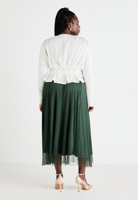 Top peplum bianco con dettagli a ruches abbinato a una gonna plissettata verde. Il modello indossa scarpe con tacco nudo, che mostrano un design testurizzato.