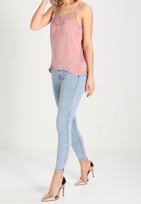 Donna che indossa un top in pizzo rosa, jeans skinny blu chiaro e scarpe con tacco alto metalliche color rosa oro, in piedi su uno sfondo semplice.
