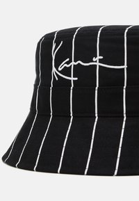 Sort bøttehatt med hvite vertikale striper, laget av slitesterkt stoff, med brodert signaturdetalj foran.