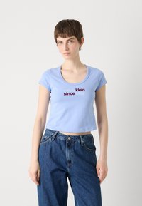 Light blue cropped t-shirt with a scoop neckline and short sleeves, featuring the text "calvin klein since 1968" in contrasting dark red. Denim jeans.