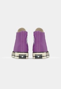 Purple canvas high-top sneakers with white eyelets and black rubber soles. Features Converse All Star logo on the sides and white stitching accents.